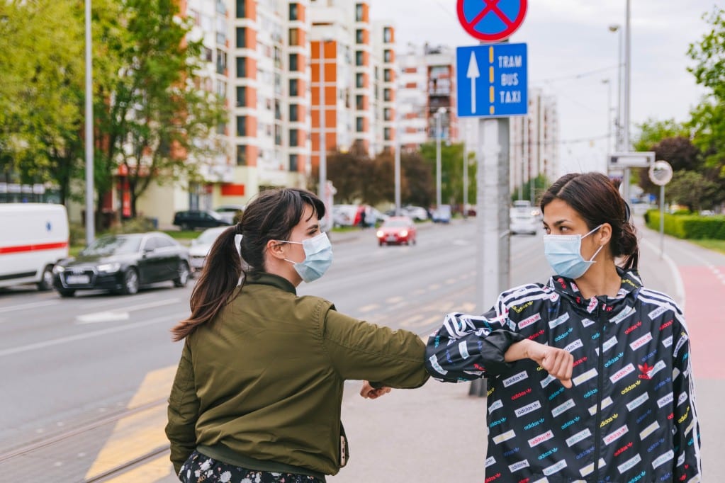 two women bump elbows on the street as they wear masks due to a COVID-19 mask mandate